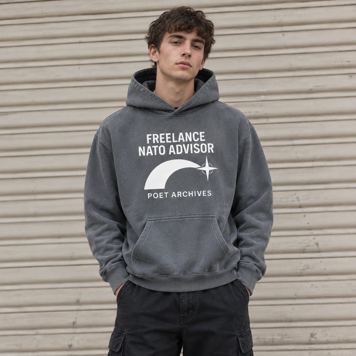 A young man wearing a dark gray washed hoodie with a white graphic print that reads 'FREELANCE NATO ADVISOR' and 'POET ARCHIVES', standing in front of a beige corrugated metal shutter.