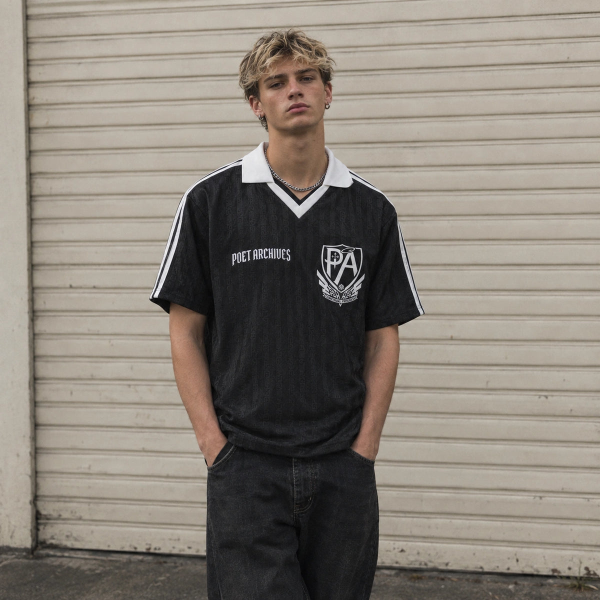 A young man wearing a black vintage-style short-sleeve t-shirt with a white collar, white sleeve stripes, and white 'POET ARCHIVES' text on the chest, standing in front of a beige corrugated metal garage door.