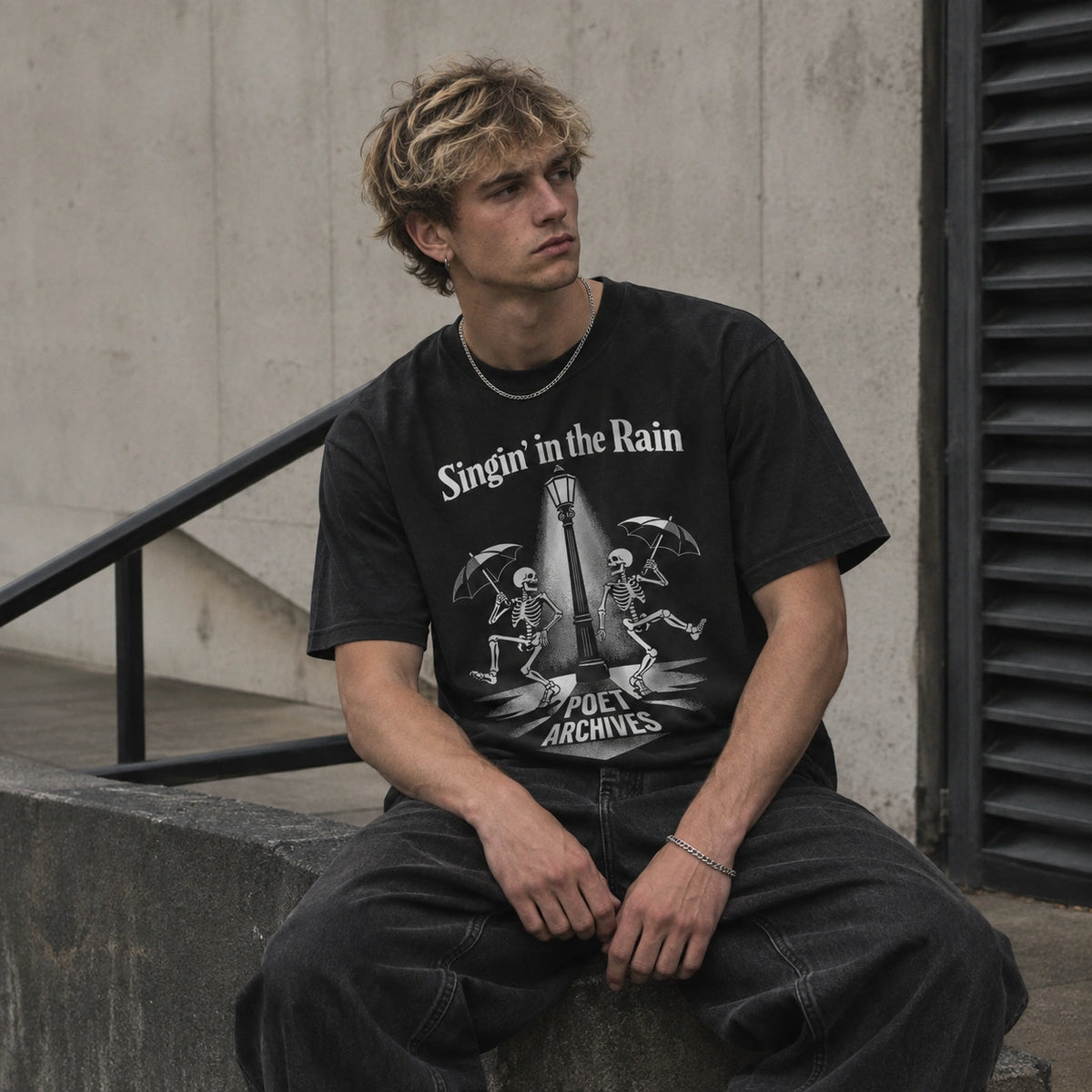 A young man with blonde hair sits on a concrete ledge wearing a black graphic t-shirt with 'Singin' in the Rain' and 'Poet Archives' printed in white, set against a concrete wall background.