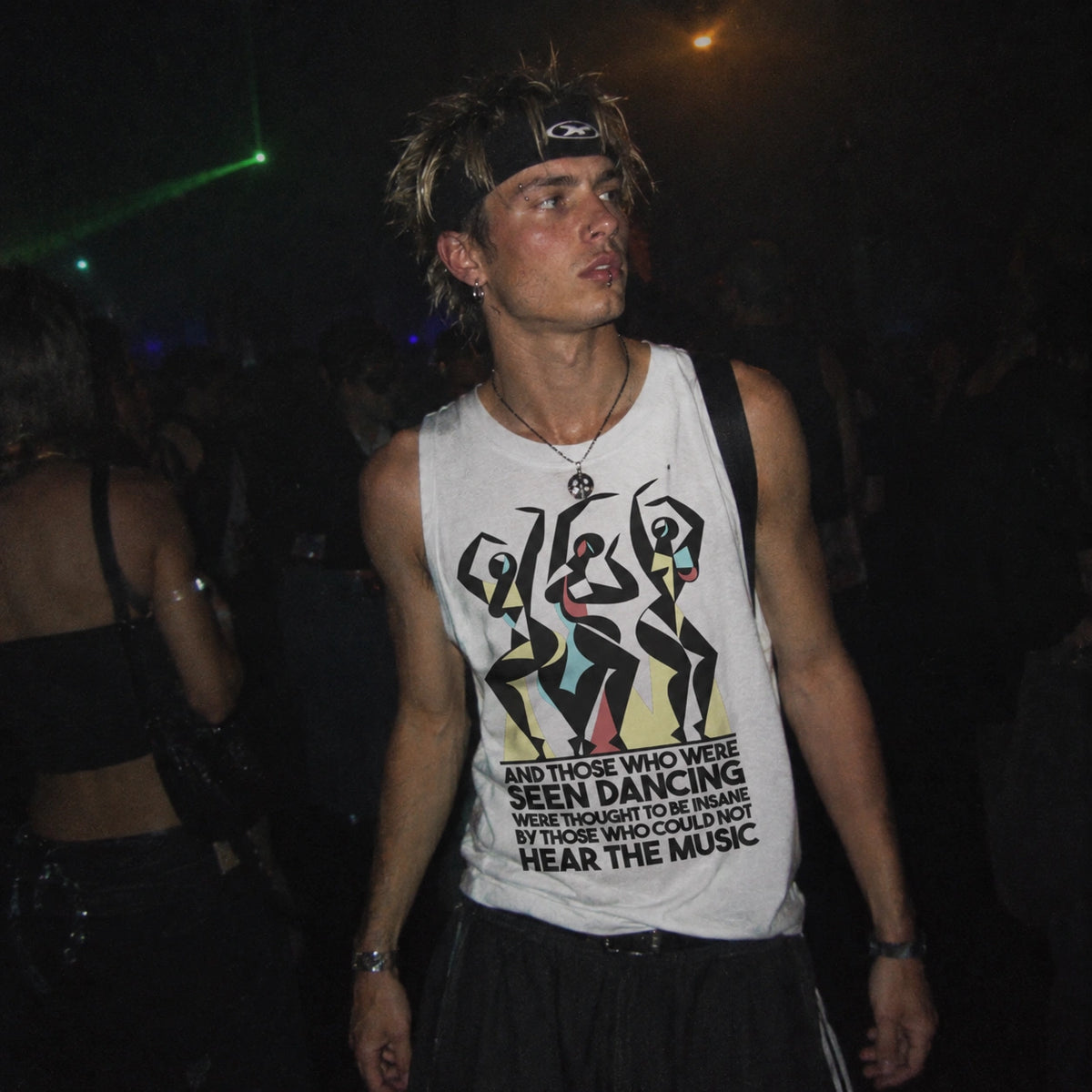A man wearing a white graphic tank top with a black and multicolor print, standing in a dark nightclub environment. The tank top features a graphic of dancing figures and the text 'AND THOSE WHO WERE SEEN DANCING WERE THOSE WHO CANNOT HEAR THE MUSIC'.