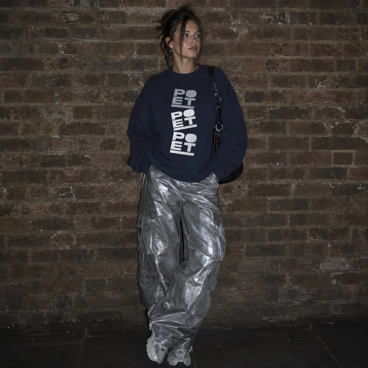 A person stands against a dark brick wall wearing an oversized navy sweatshirt with large white vertical text, shiny metallic silver pants, and white sneakers, with a black shoulder bag.