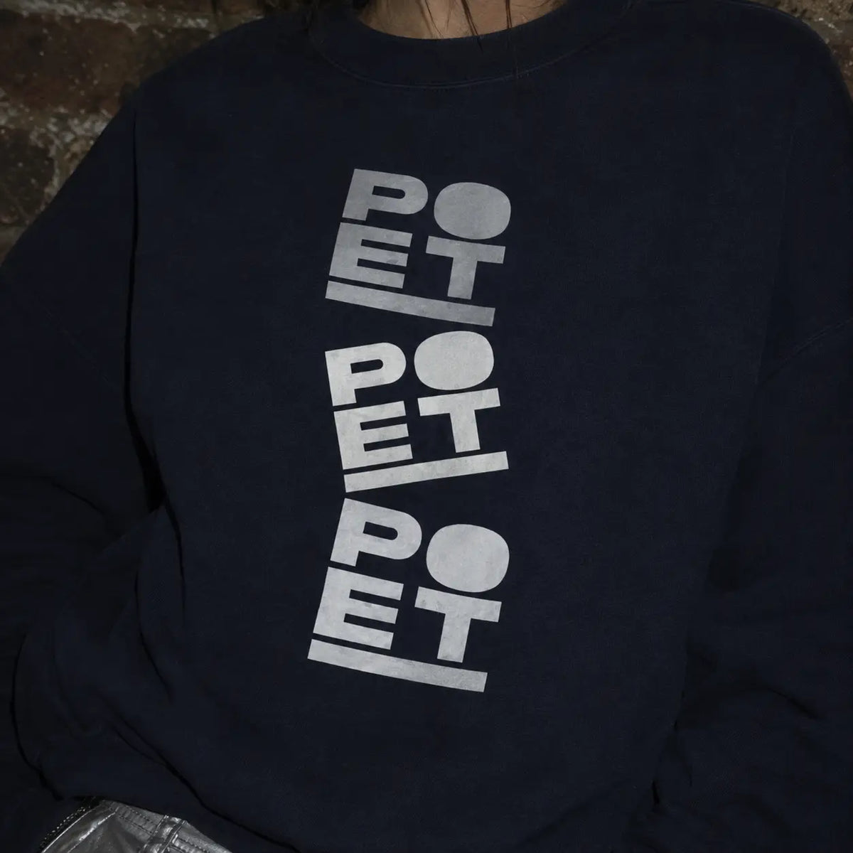 A close-up of a person wearing a dark navy crewneck sweatshirt with a vertical graphic print on the front. The graphic features the word 'POET' repeated three times in a stacked, stylized design using white, gray, and light blue-gray blocks.
