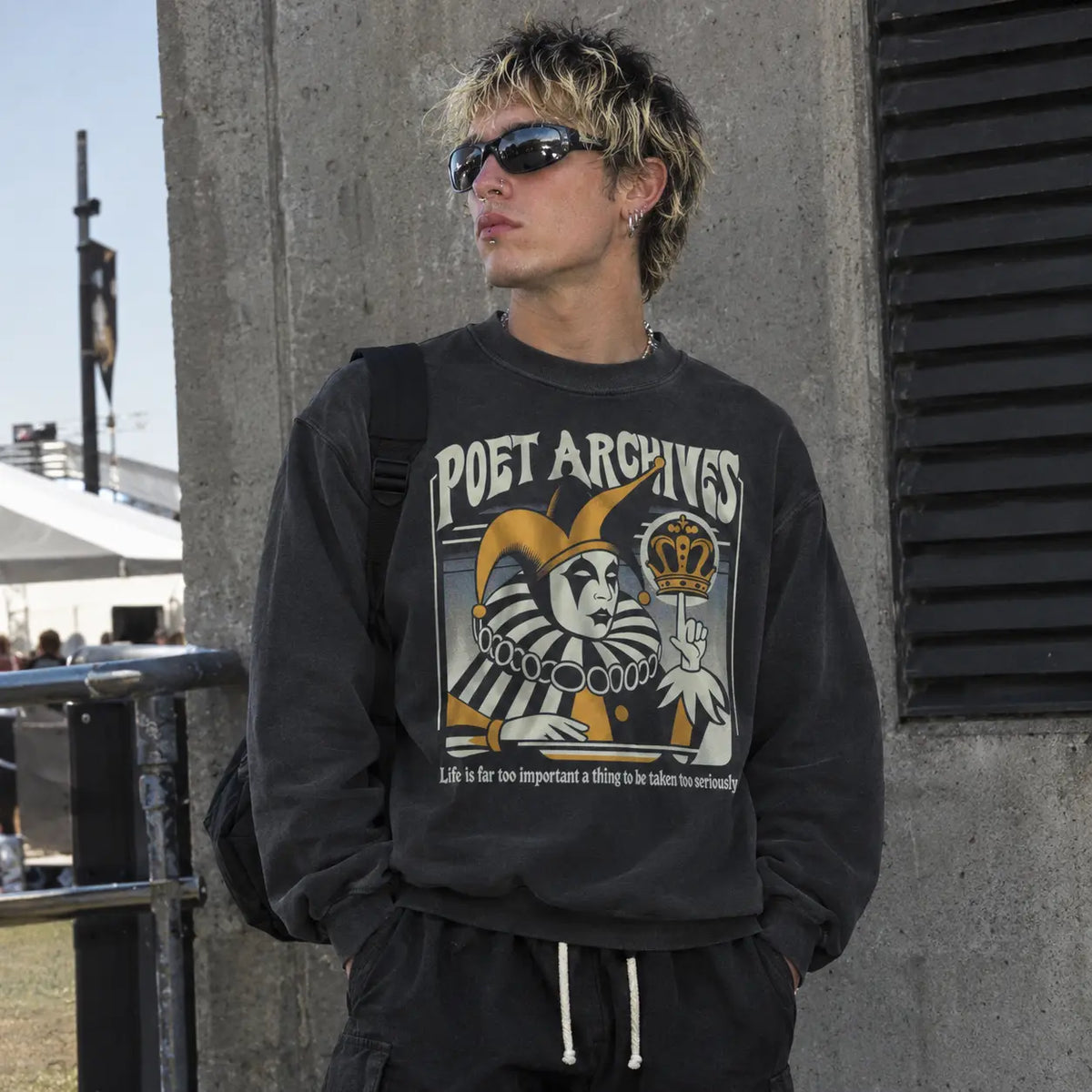 A man wearing a black graphic sweatshirt with 'POET ARCHIVES' and a jester illustration, paired with black sweatpants and sunglasses, standing outdoors next to a concrete pillar.