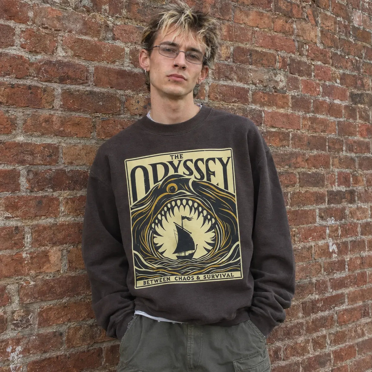 A young man with glasses and blonde hair stands against a red brick wall wearing a dark brown crewneck sweatshirt with a large graphic print of a shark and the text 'THE ODYSSEY' and 'BETWEEN CHAOS & SURVIVAL'.