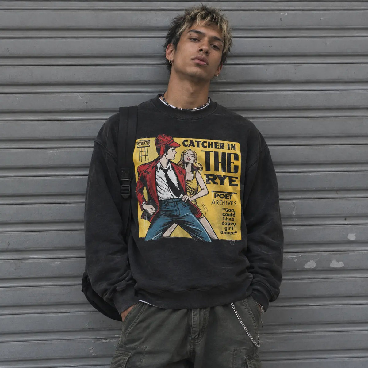 A young man wearing a washed black crewneck sweatshirt with a vintage-style graphic print of a man in a red jacket and blue jeans, leaning against a gray metal roll-up door.