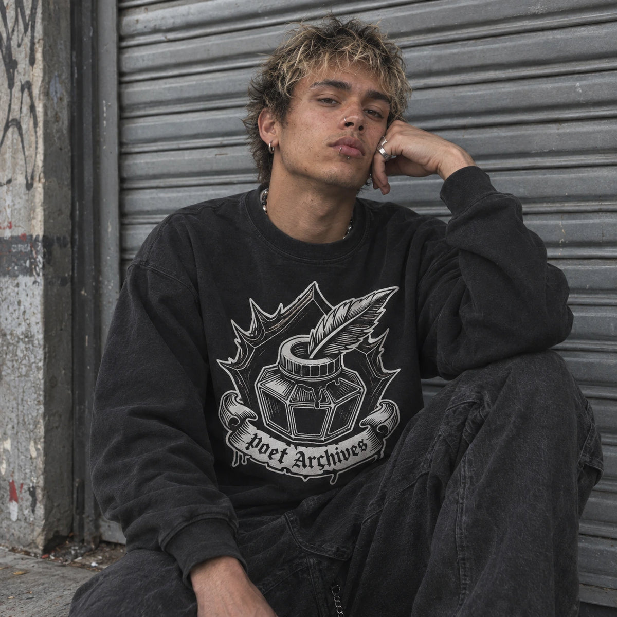 A young man with blonde-highlighted hair sits in front of a gray metal shutter, wearing a washed black crewneck sweatshirt with a white graphic of an ink bottle and quill with the text 'poet Archives'.