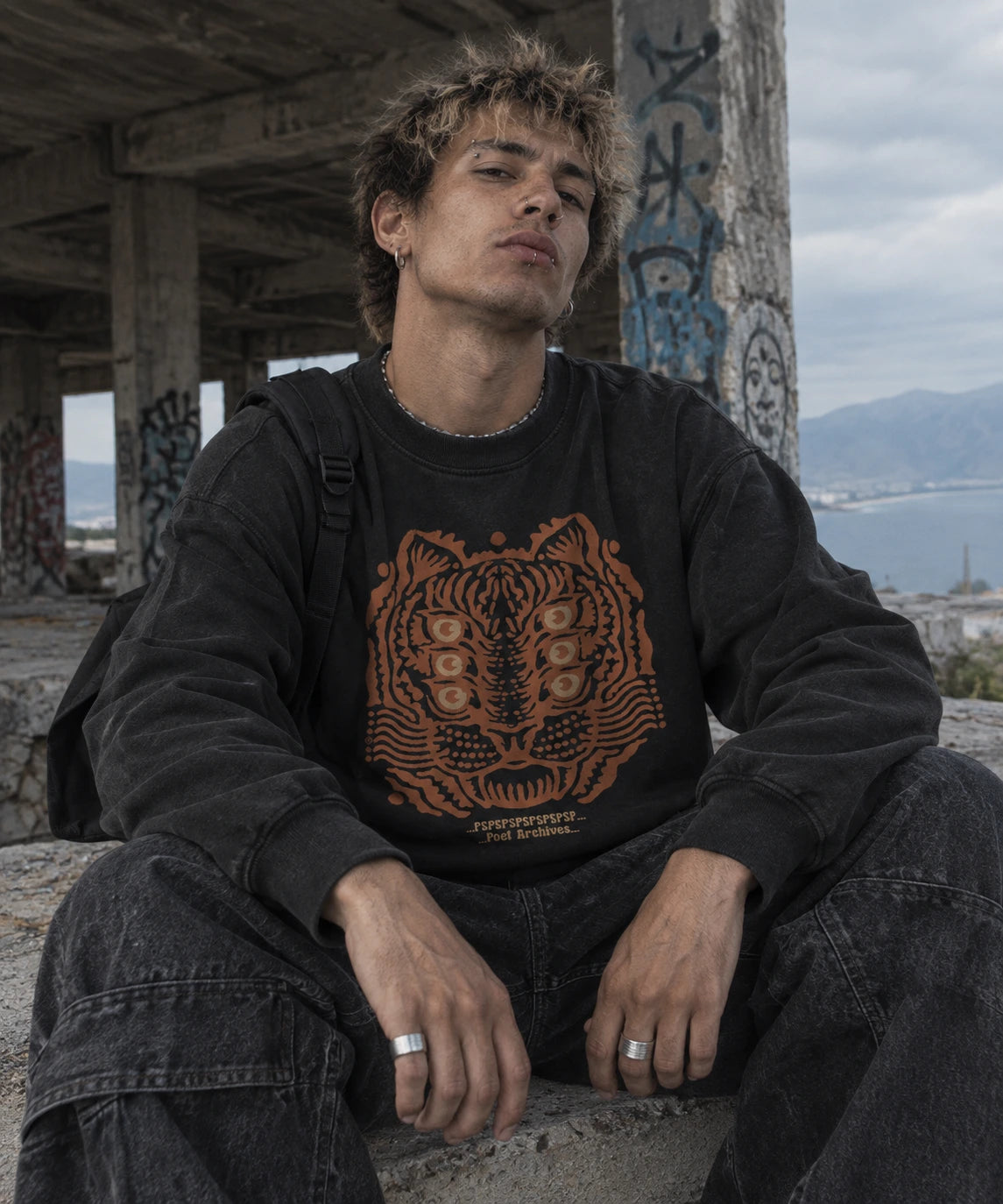 A young man sitting outdoors in a concrete structure, wearing a black crewneck sweatshirt with a large orange tiger graphic and the brand name 'POET ARCHIVES' printed below it, paired with dark jeans.