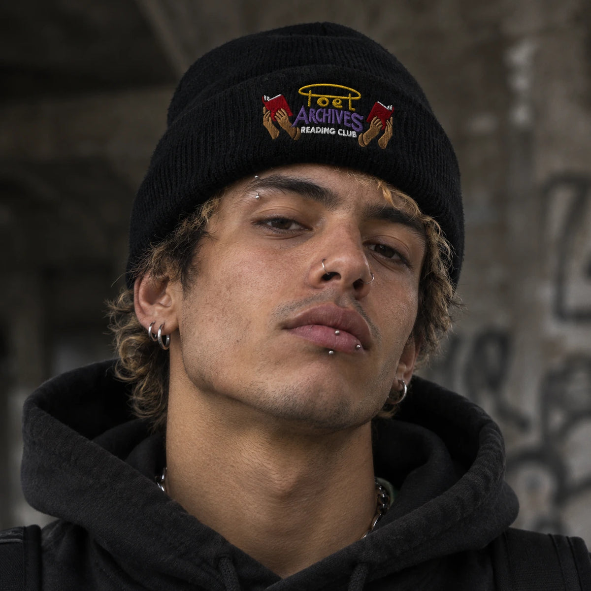 A close-up portrait of a young man wearing a black knit beanie with a colorful embroidered logo that reads 'Poet Archives Reading Club'.