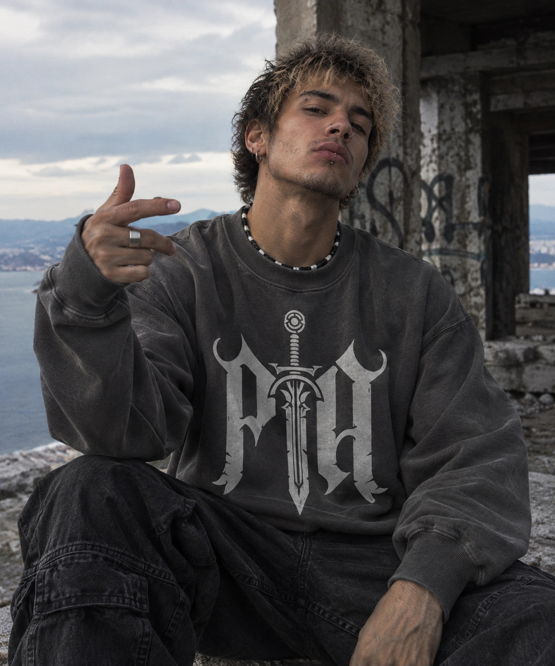 A man wearing a dark gray washed sweatshirt with a white gothic letter 'PHA' and sword graphic, sitting in an outdoor ruin with water and sky in the background.