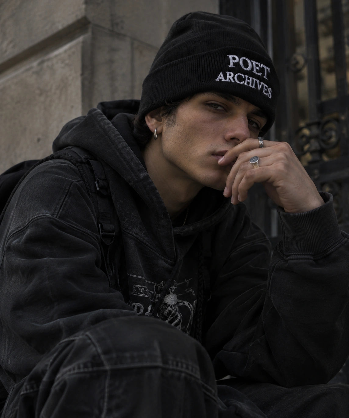 A man wearing a black beanie with 'POET ARCHIVE' in white text, a black washed hoodie, and a backpack, sitting against a stone wall.