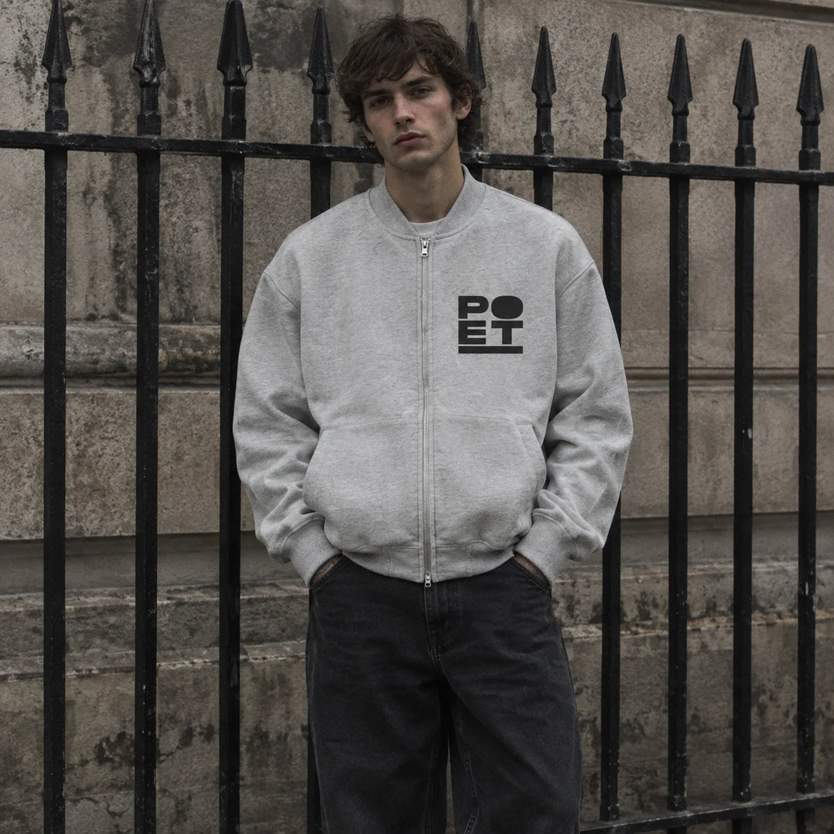 A man wearing a light gray zip-up jacket with a black 'POET' logo on the chest, standing in front of a black wrought-iron fence and stone wall.
