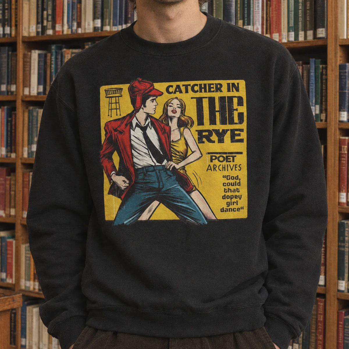 A person wearing a black crewneck sweatshirt with a vintage-style comic book graphic of a man and woman in front of a yellow poster reading 'CATCHER IN THE RYE' and 'POET ARCHIVES'. The background consists of wooden bookshelves filled with books.