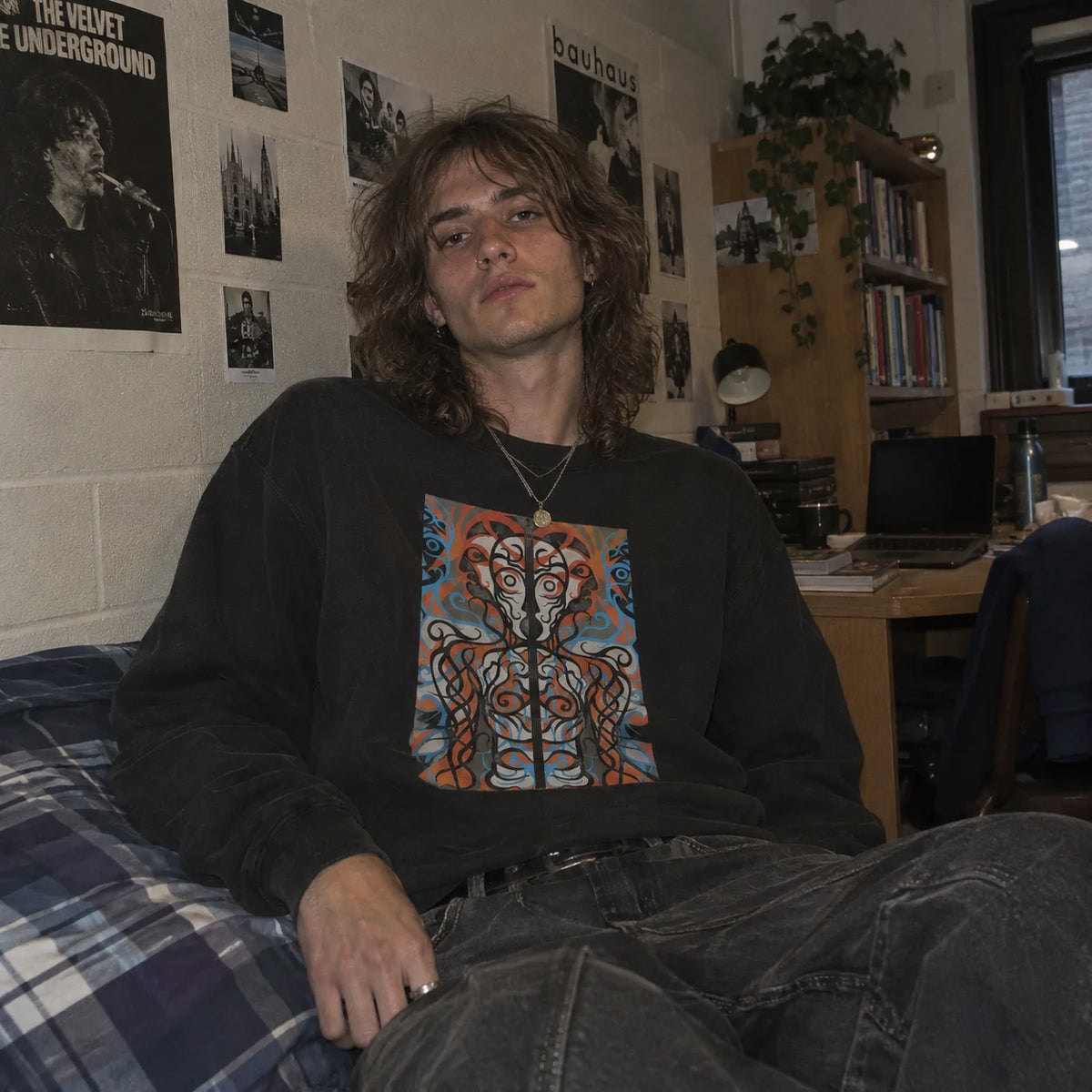 Young man with long brown hair sitting on a bed wearing the black Humanoid Sweatshirt, with Velvet Underground and Bauhaus posters and a bookshelf in the background