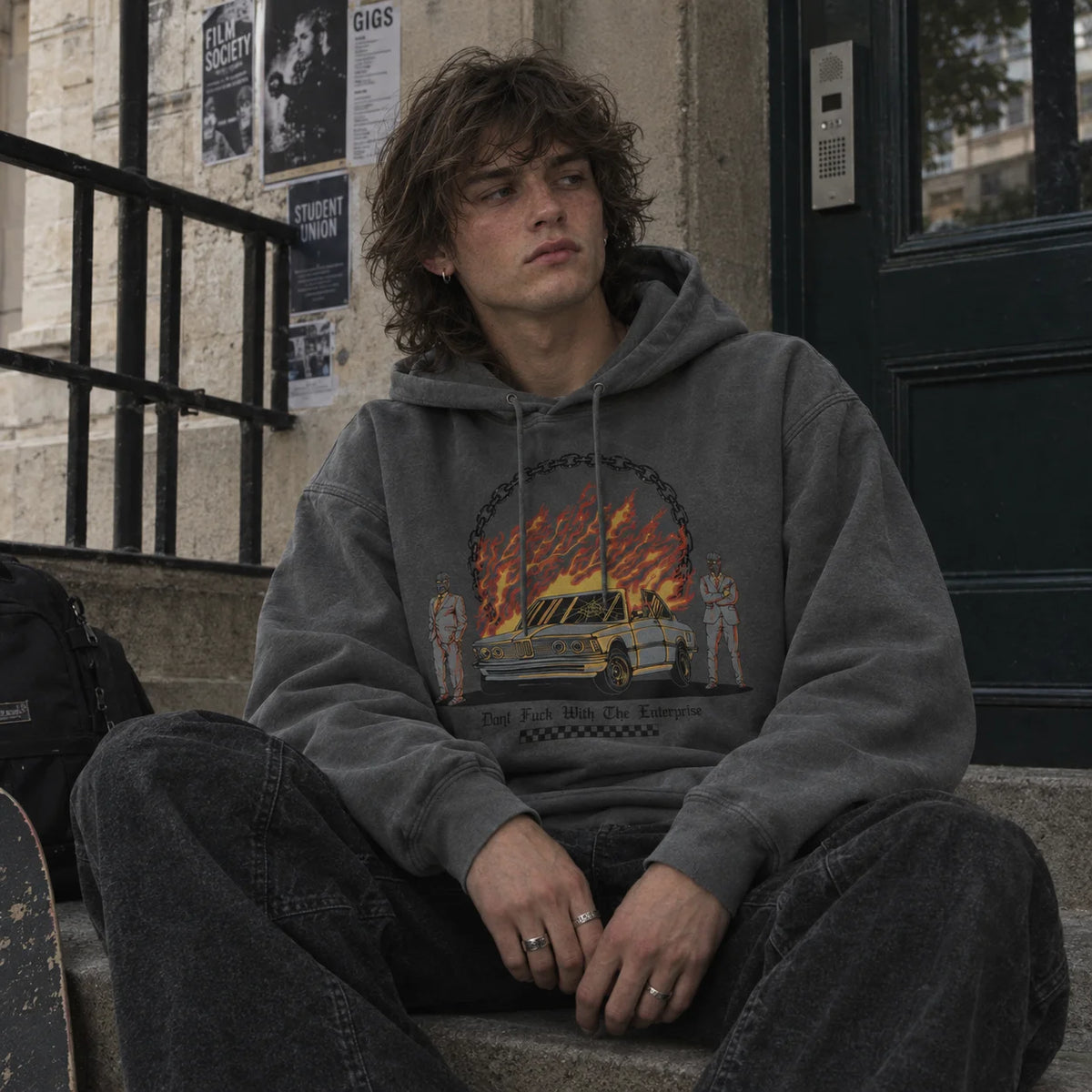 A young man with curly brown hair sits on concrete steps wearing a faded dark gray hoodie with a graphic print of a car on fire .