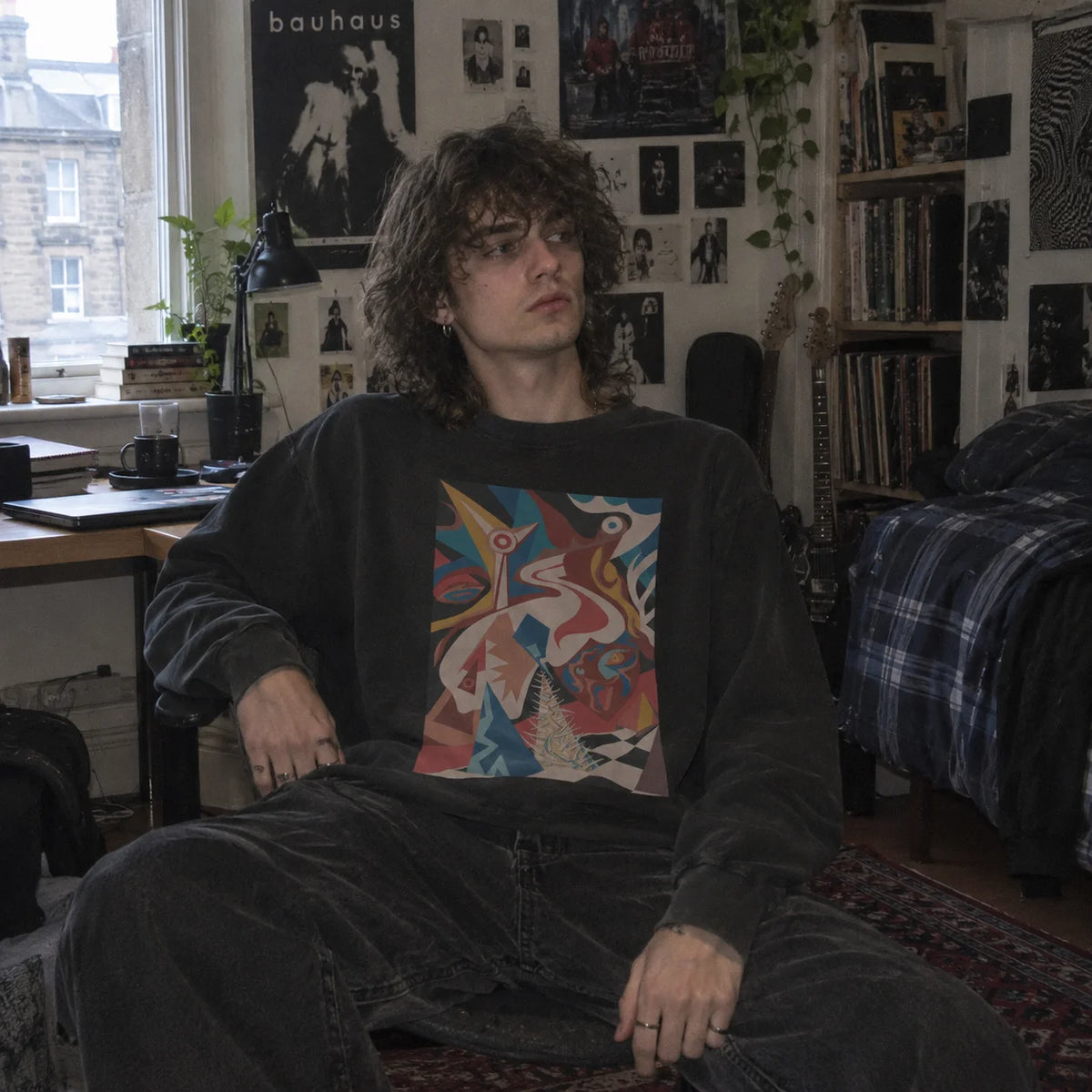 A person sitting in a dimly lit, cluttered bedroom or studio space, wearing a faded black long-sleeve crewneck sweatshirt with a colorful abstract graphic print on the front.
