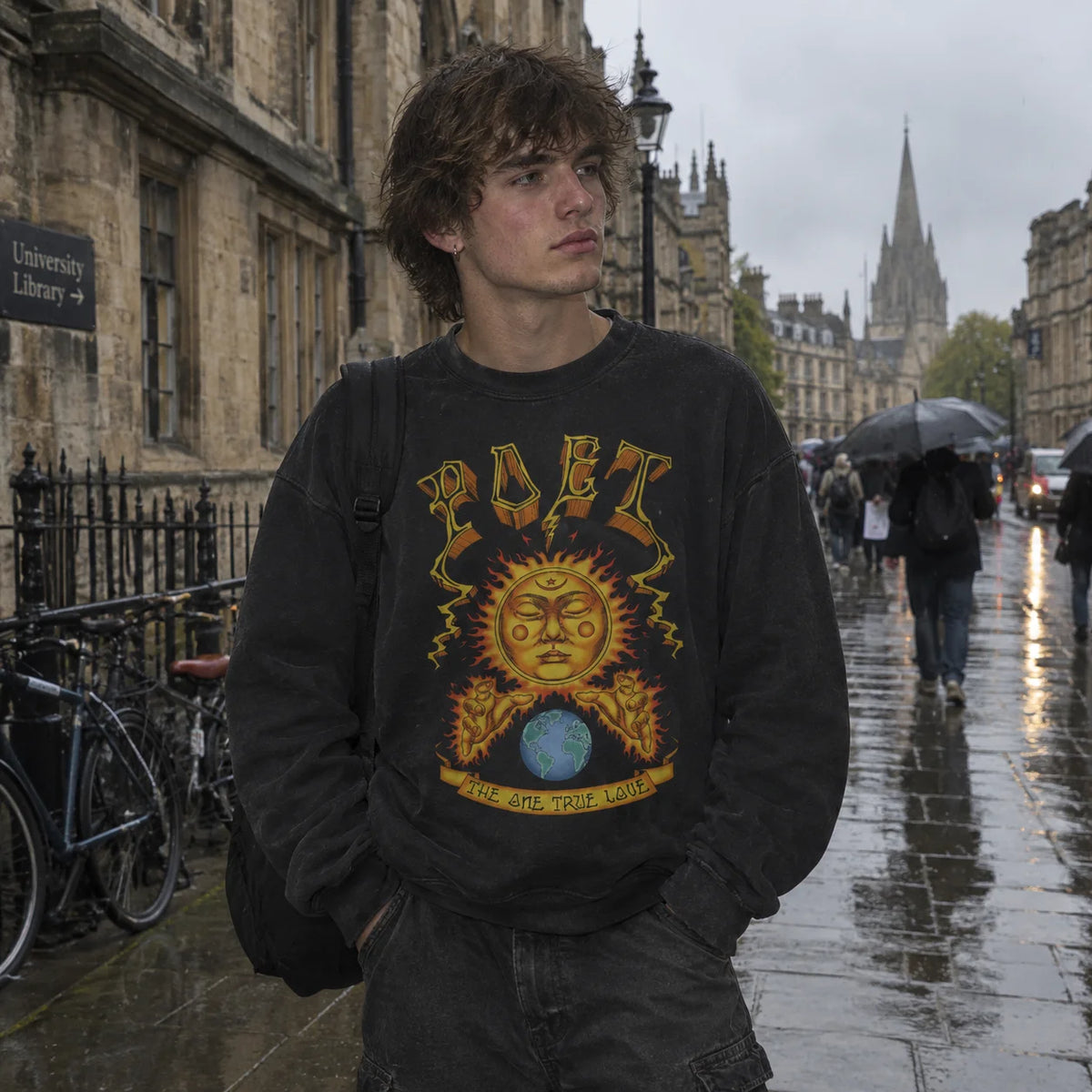 A young man standing on a wet city street wearing a faded black crewneck sweatshirt with a large yellow, orange, and blue graphic print on the front. The background shows historic stone buildings, a rainy street scene with pedestrians, and a church spire in the distance.