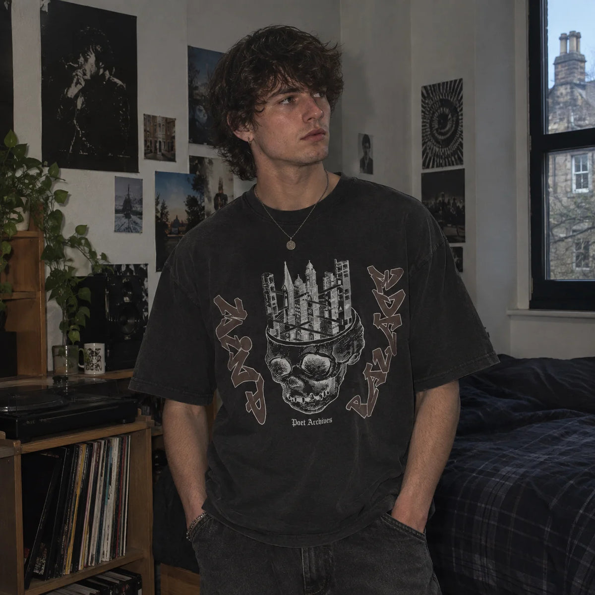 A man wearing a faded black graphic t-shirt with a skull and cityscape design, standing in a bedroom with posters on the wall.