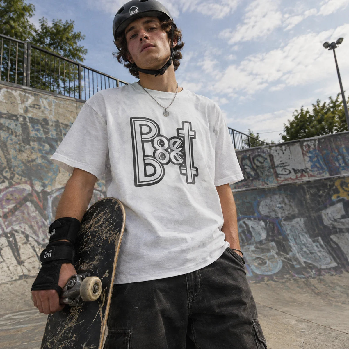 Man at a skatepark holding a skateboard, wearing a white t-shirt with a mirrored 'Poet' logo graphic