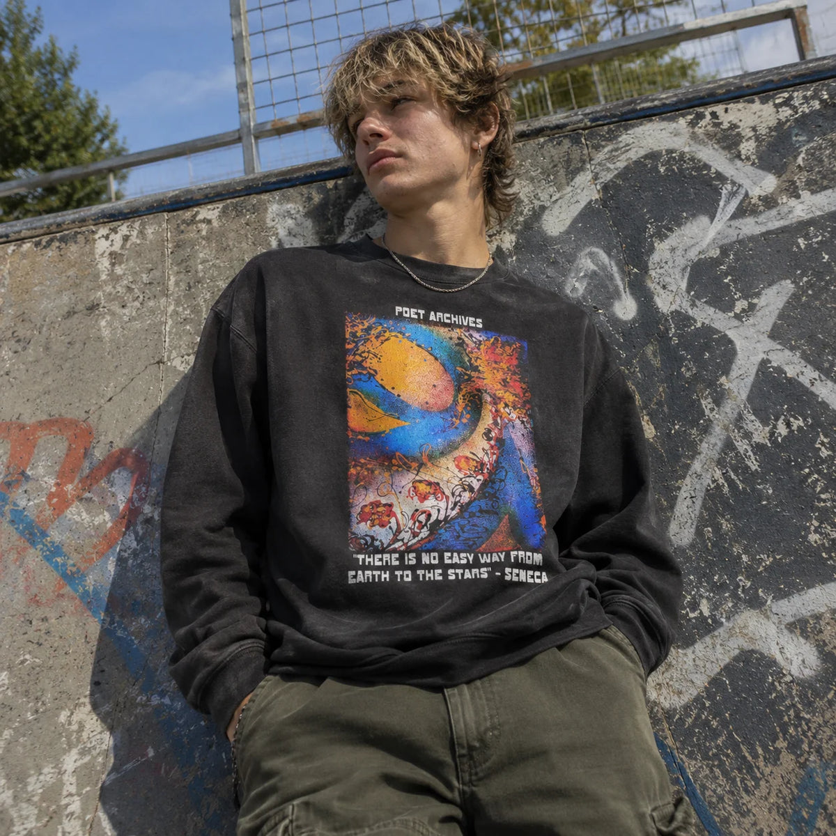 A young man with blonde hair leans against a concrete skatepark wall wearing a black graphic sweatshirt with a colorful abstract print and the text 'POET ARCHIVES' and 'THERE IS NO EASY WAY FROM EARTH TO THE STARS - SENECA'.