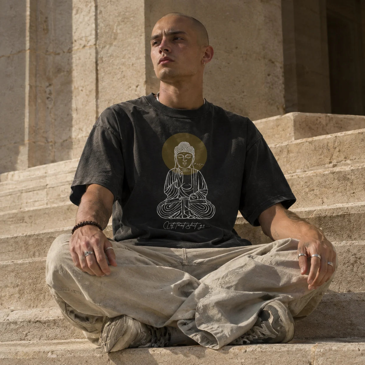 A man sits cross-legged on stone steps wearing a faded black graphic t-shirt with a white Buddha illustration and gold circle, paired with light beige pants.