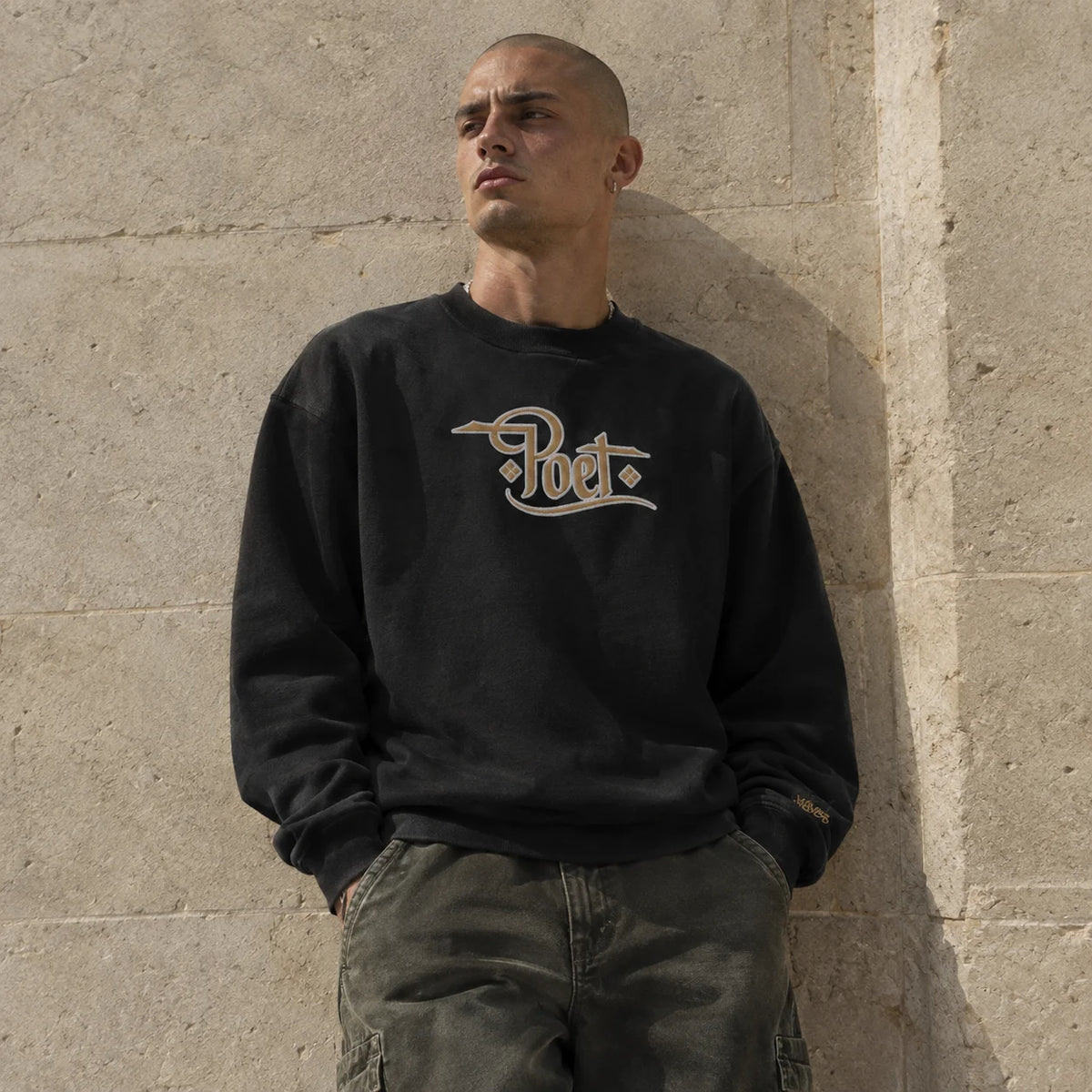 A man wearing a black crewneck sweatshirt with a beige 'Poet.' logo on the chest, leaning against a textured concrete wall.