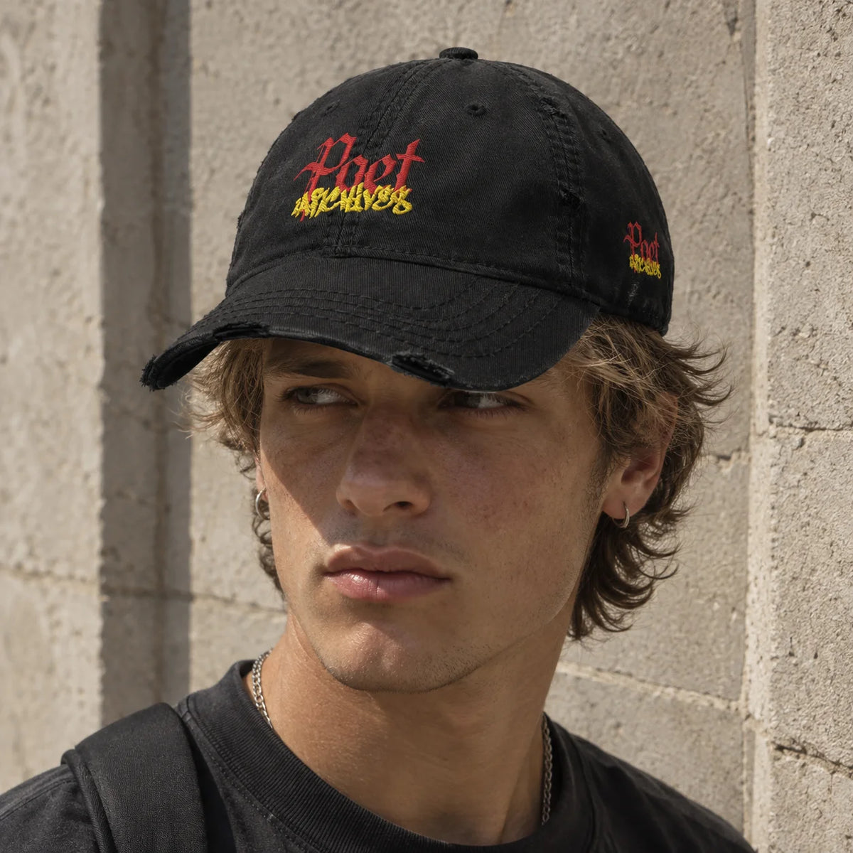 A man wearing a black distressed baseball cap with red and yellow embroidered text reading  "Poet Archives" against a light concrete wall.