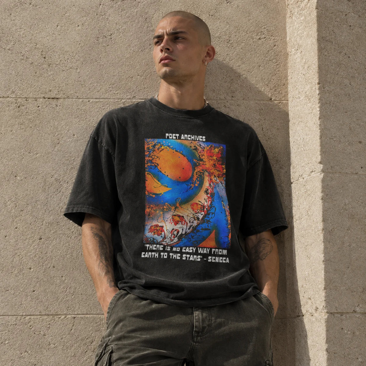 A man wearing a faded black graphic t-shirt with a colorful abstract print and text, paired with dark cargo pants, leaning against a textured concrete wall.