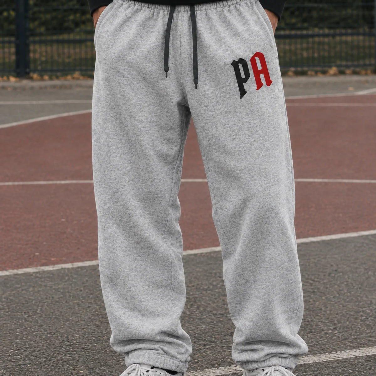 A person wearing light heather gray jogger sweatpants with a black and red 'PA' logo on the left thigh, standing on an outdoor basketball court.