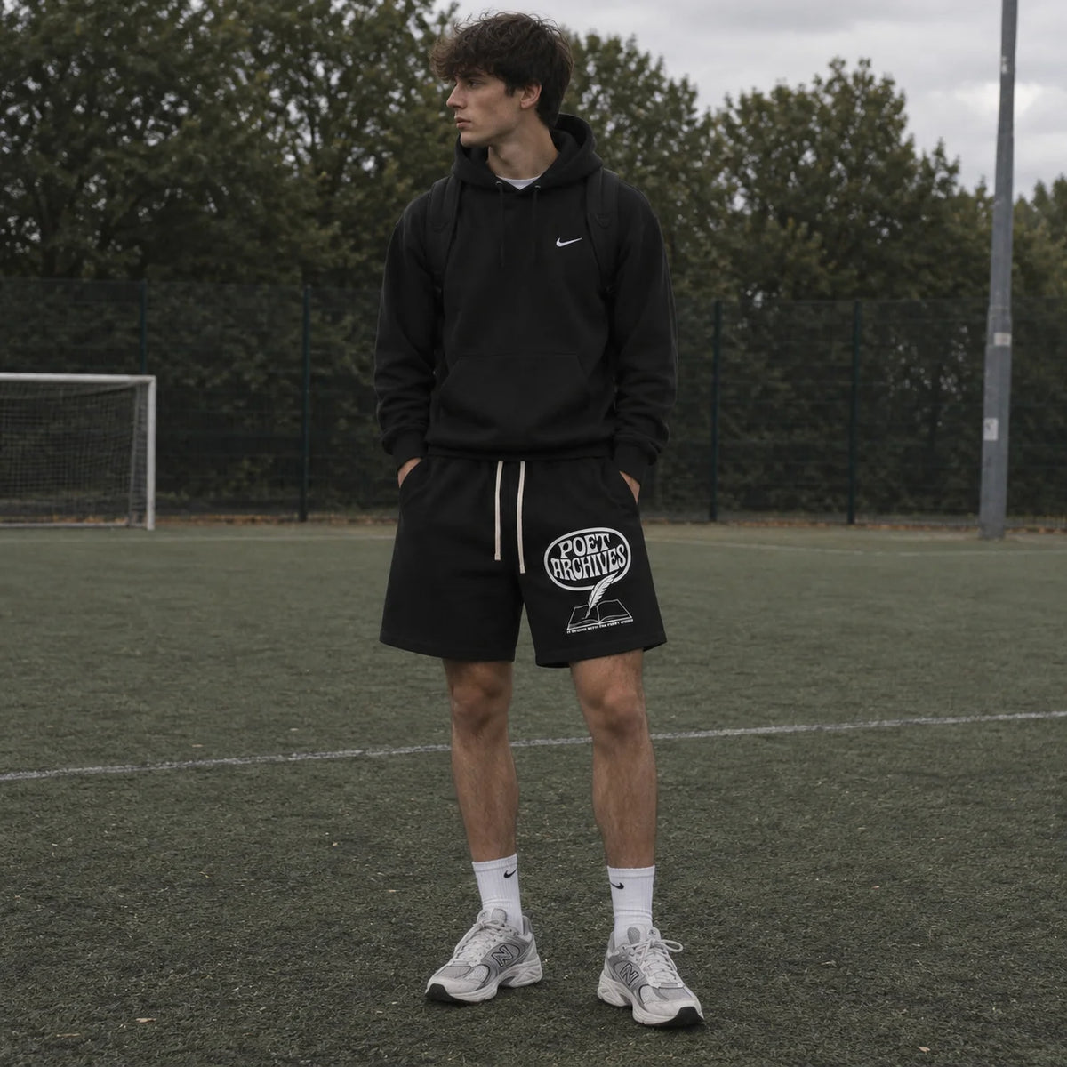A man wearing a black hoodie and the First Word Shorts with a white graphic logo, on an outdoor sports field