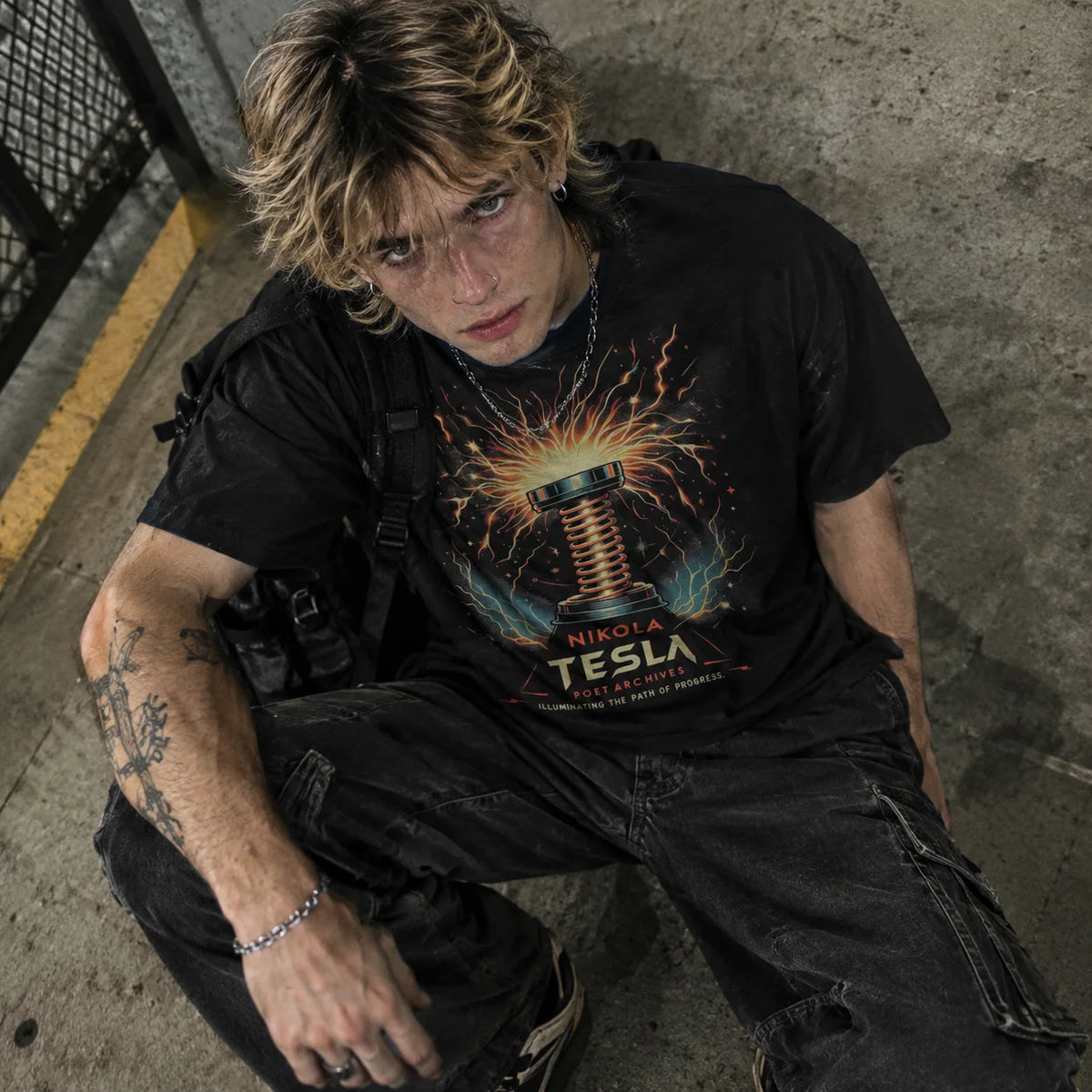 A man wearing a black graphic t-shirt with a colorful Tesla coil design and the text 'Nikola Tesla' and 'Inventor of Modern Electricity', paired with black cargo pants, sitting against a concrete wall.