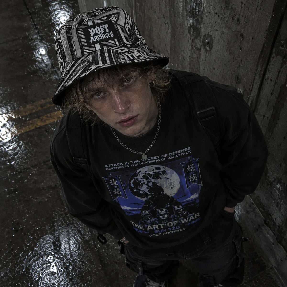 A person wearing a black graphic t-shirt and a black-and-white patterned bucket hat, looking down in a dark, wet alleyway. The t-shirt features a blue and black graphic with the text 'ATTACK IS THE SECRET OF DEFENSE DEFENSE IS THE SECRET OF ATTACK THE ART OF WAR' and 'PORT AIRPLANE'.