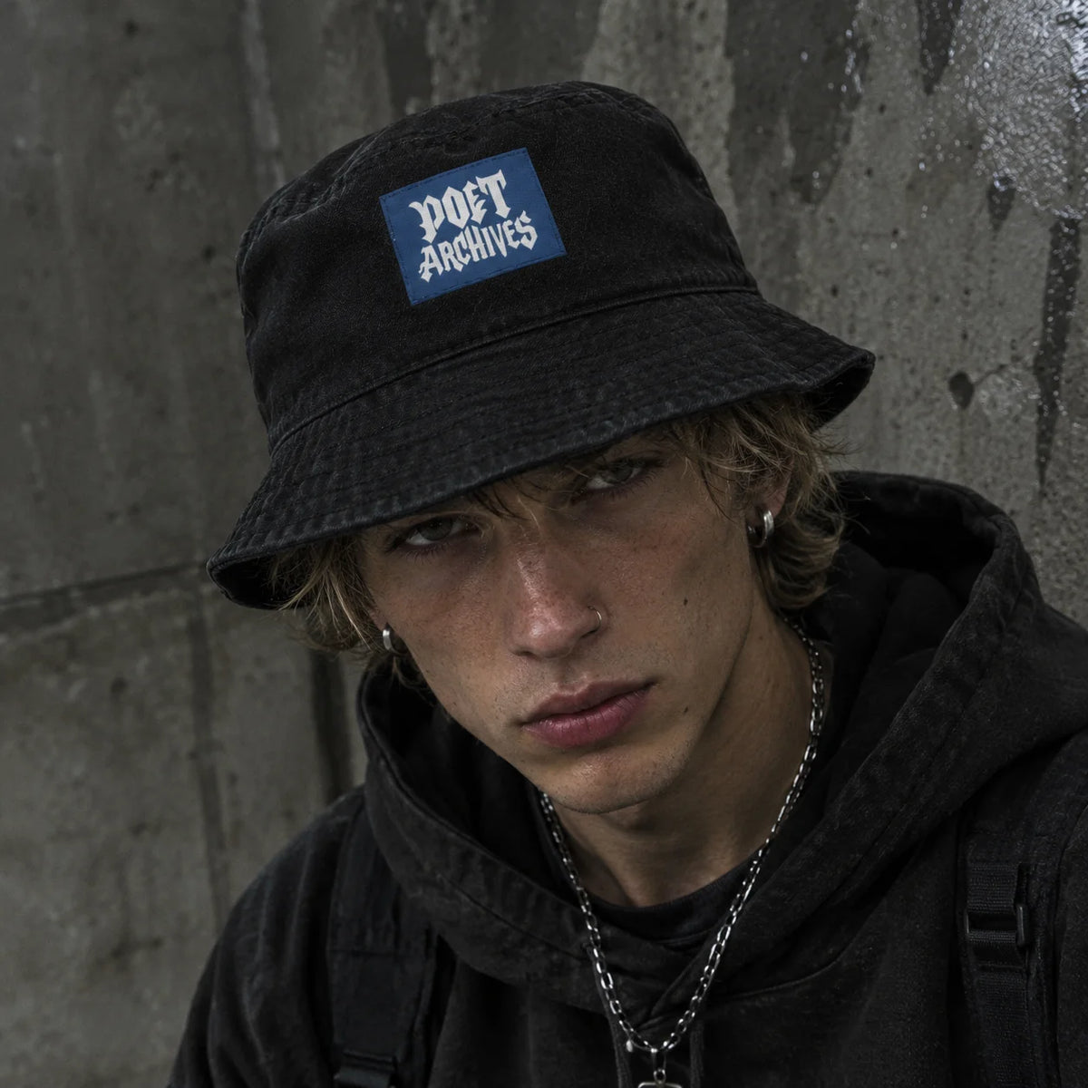 A young man wearing a black bucket hat with a blue 'POET ARCHIVES' patch on the front, set against a textured concrete wall.