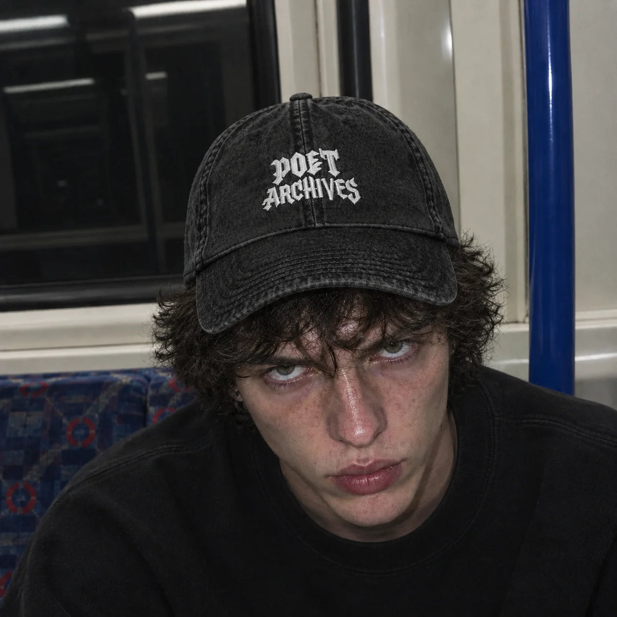 A person wearing a black washed baseball cap with 'POET ARCHIVES' embroidered in white gothic lettering, photographed in a real indoor environment with a window and blue pole visible in the background.