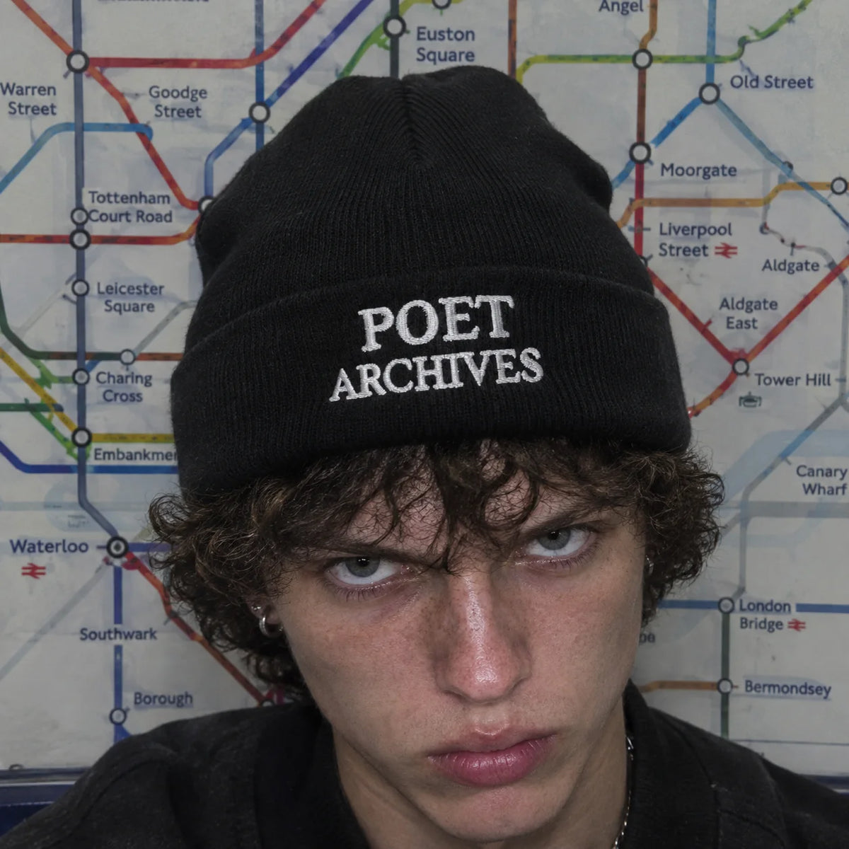 A close-up of a person wearing a black knit beanie with 'POET ARCHIVES' embroidered in white on the front, set against a London Underground map background.