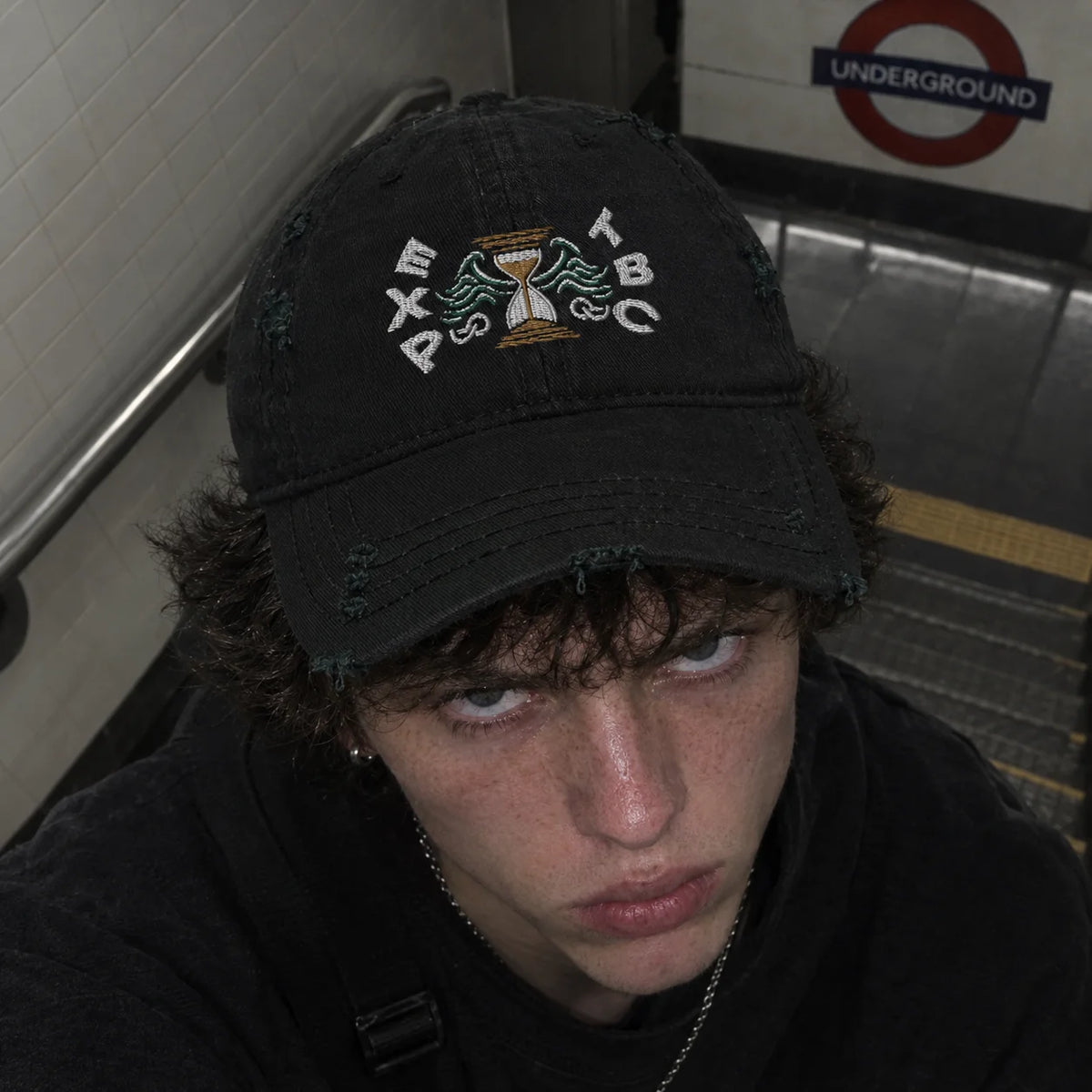 A close-up shot of a person wearing a black distressed baseball cap with white embroidered text and a central graphic, photographed in a subway station.