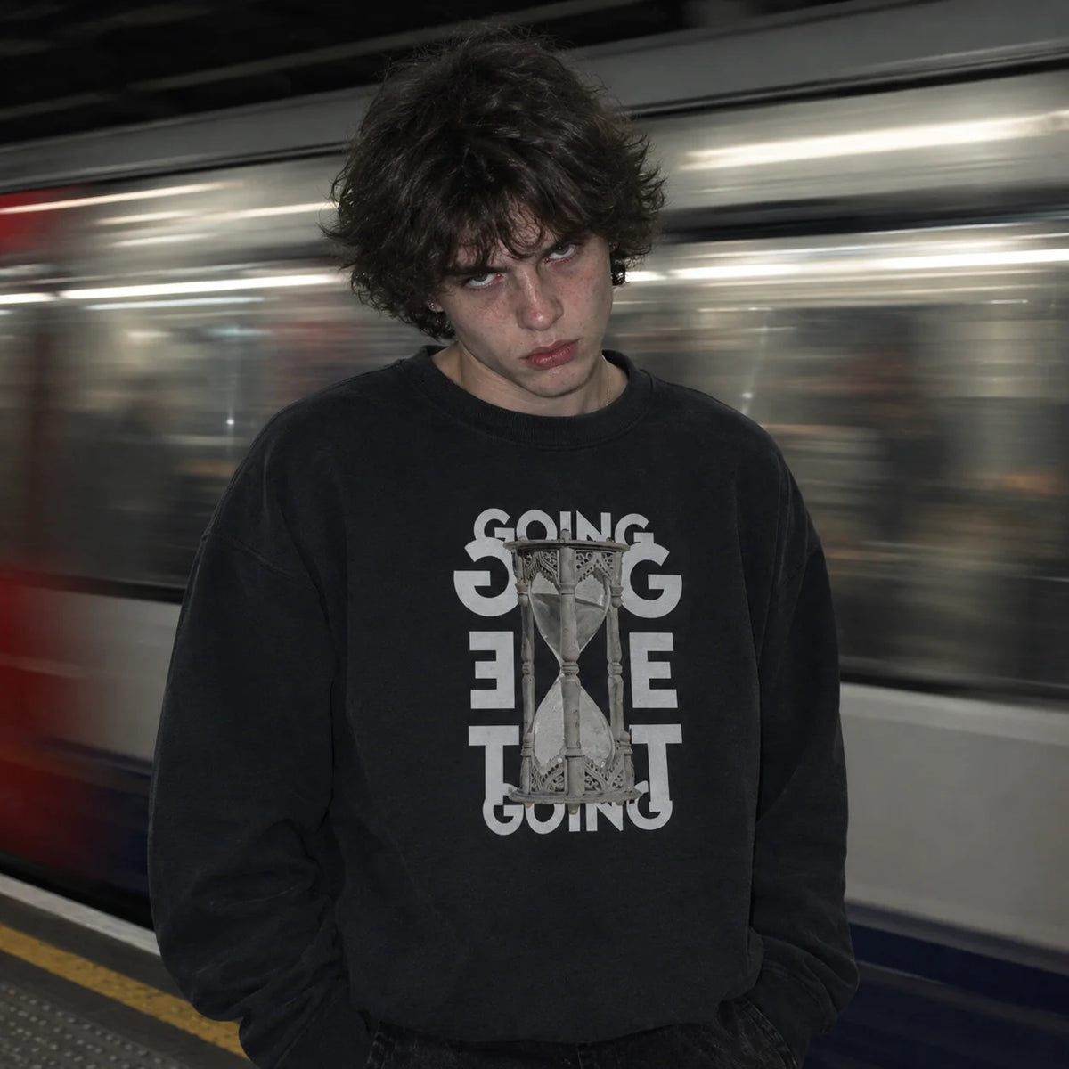 A person standing on a train platform wearing a black crewneck sweatshirt with a white graphic print that reads 'GOING GET TO GOING' around an hourglass design. A blurred train is visible in the background.
