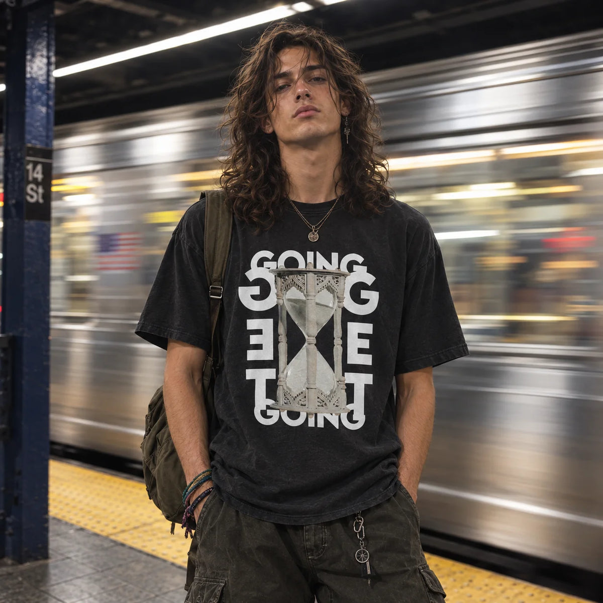 A man with long curly hair stands on a subway platform wearing a black graphic t-shirt with a white and gray hourglass design and the words 'GOING GET TO GOING'. A blurred silver train passes in the background of a subway station.