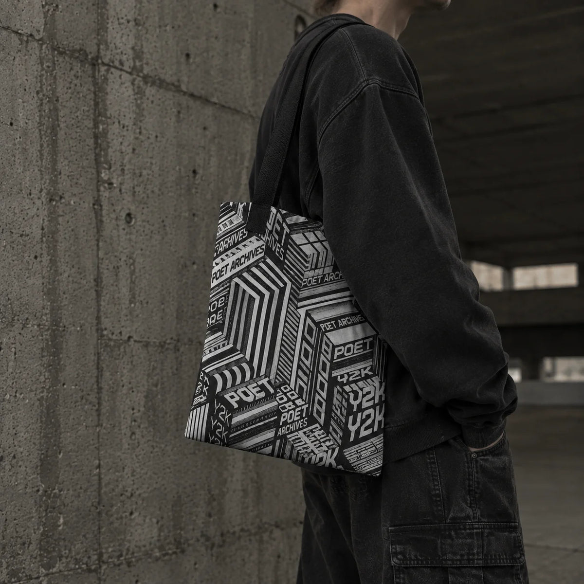 A person wearing a black long-sleeve shirt and dark cargo pants carries a black and white geometric patterned tote bag over their shoulder while standing against a concrete wall.
