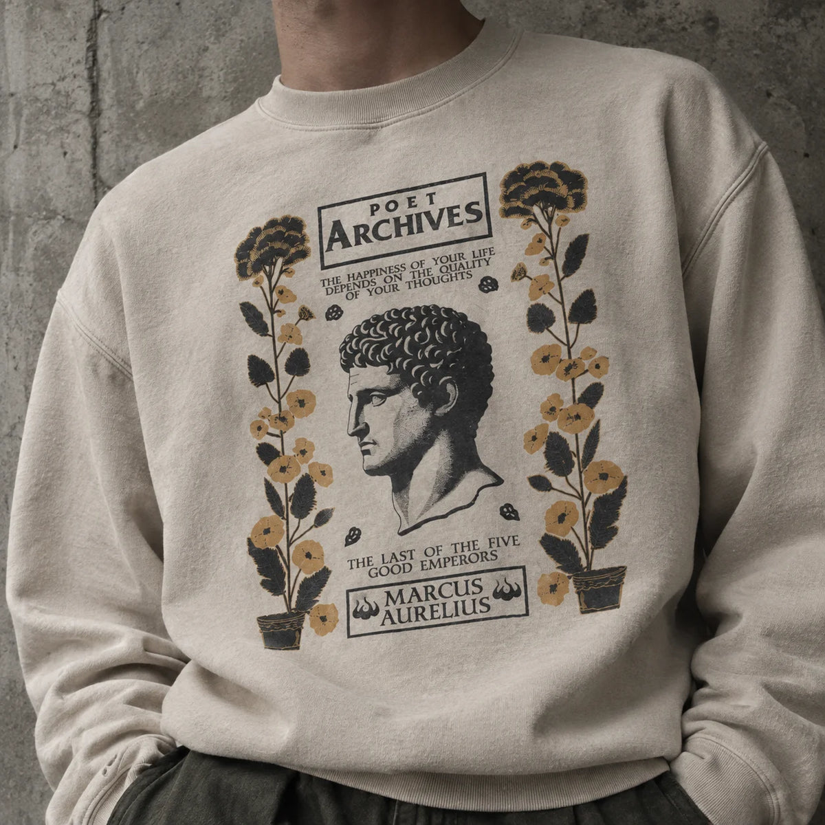 A person wearing a cream-colored crewneck sweatshirt with a vintage-style graphic print featuring a portrait of Marcus Aurelius and the text 'Poet Archives' and 'Marcus Aurelius'. The background is a textured gray concrete wall.