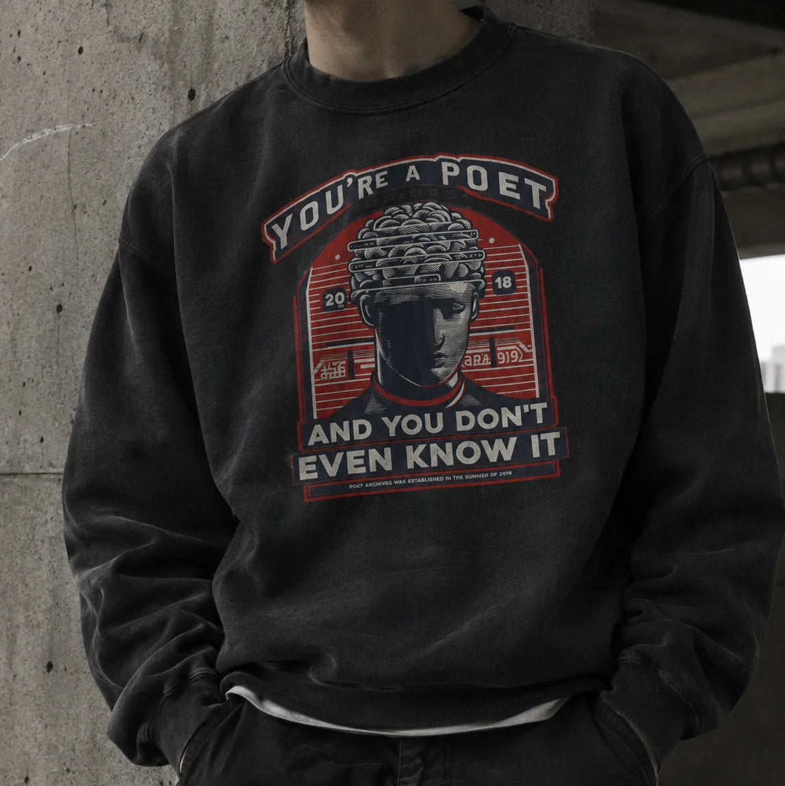 A person wearing a dark charcoal gray crewneck sweatshirt with a vintage-style graphic print featuring a classical statue and the text 'YOU'RE A POET AND YOU DON'T EVEN KNOW IT' in red, white, and gray.