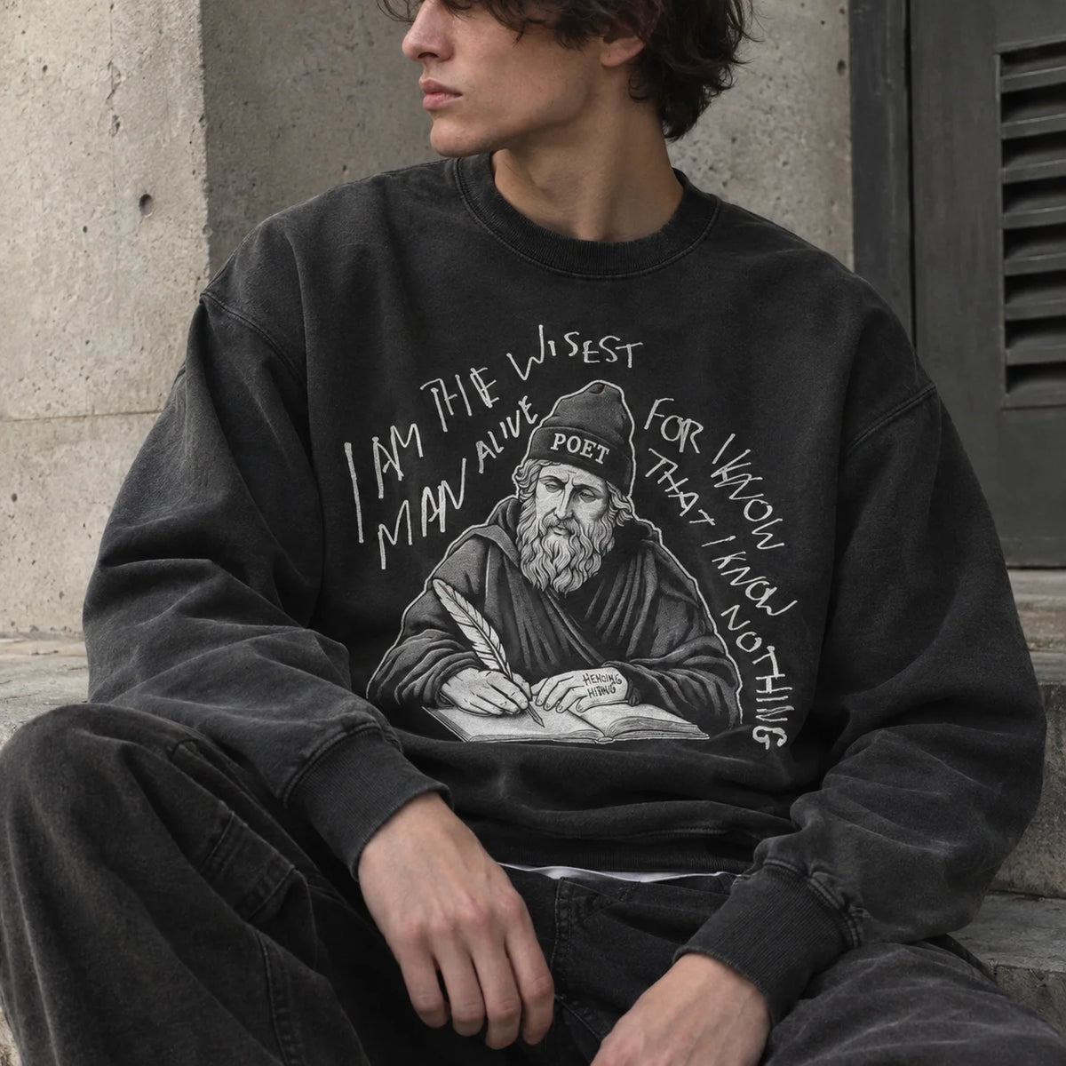 A person wearing a faded black crewneck sweatshirt with a white graphic print of a bearded poet and handwritten text, seated against a concrete wall.
