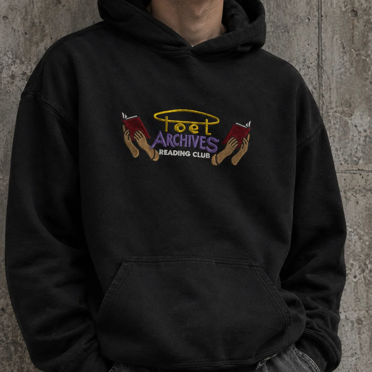 A person wearing a black pullover hoodie with a colorful graphic on the chest that reads 'Poet Archives Reading Club' with two hands holding red books, posed against a textured gray concrete wall.