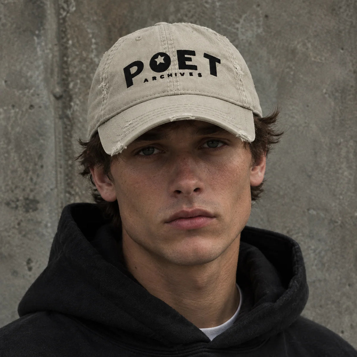 A close-up portrait of a young man wearing a distressed beige baseball cap with 'POET ARCHIVES' printed in black on the front, paired with a black hoodie against a textured gray concrete wall.
