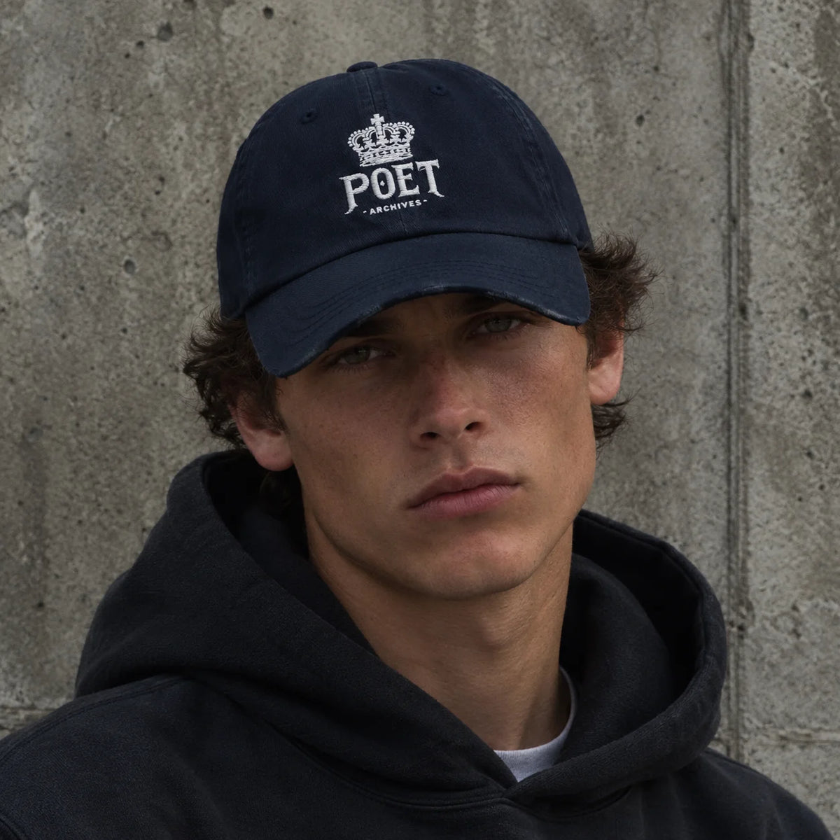 A man wearing a navy blue POET ARCHIVES baseball cap and a dark hoodie, photographed against a textured concrete wall.