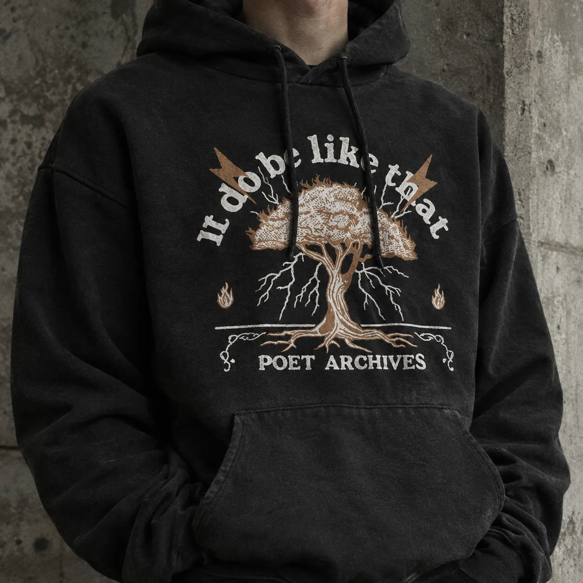 A person wearing a faded black pullover hoodie with a graphic print of a tree, flames, and the text 'I do b* like that' and 'POET ARCHIVES' on the front, set against a textured concrete wall.