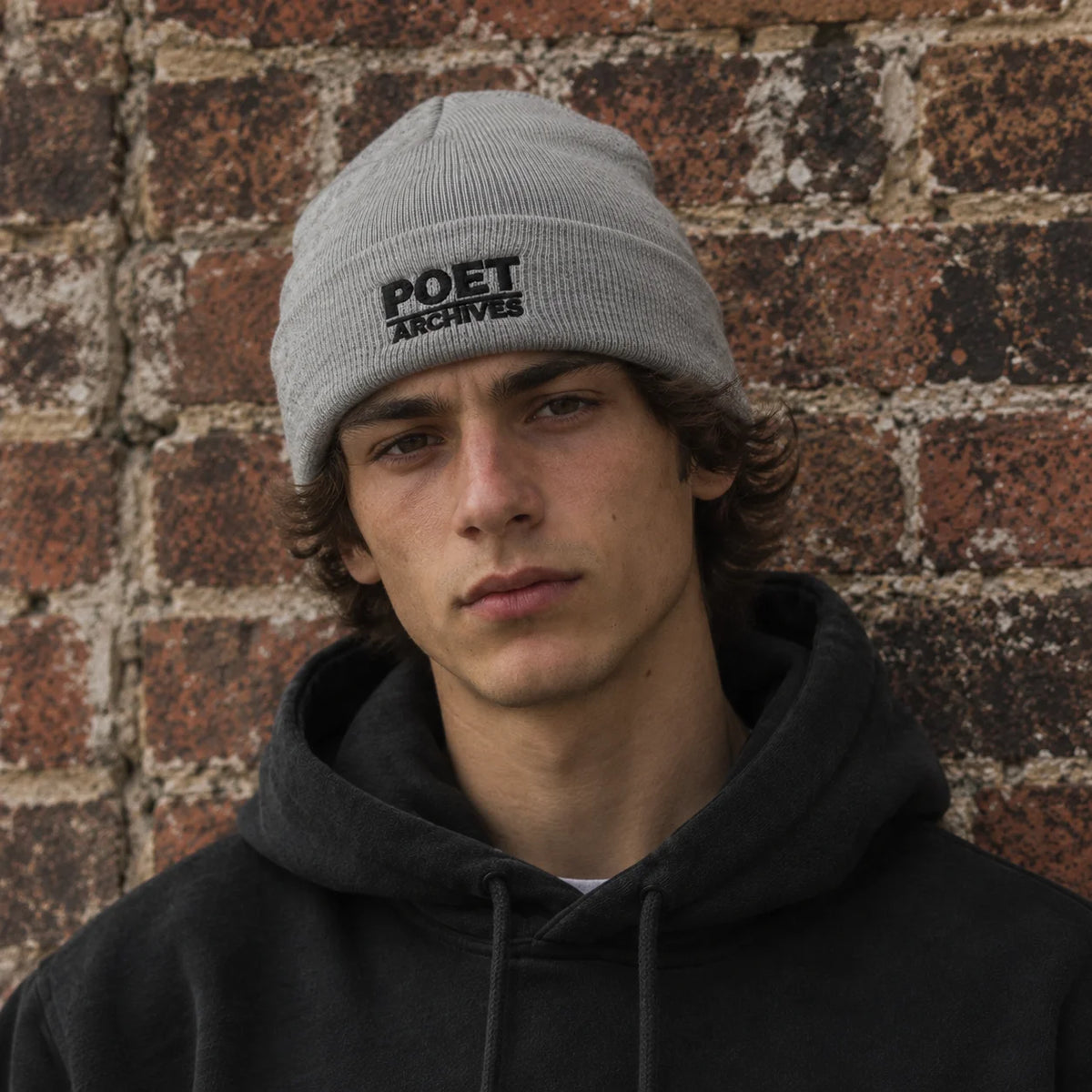 A young man wearing a light gray knit beanie with 'POET ARCHIVES' embroidered in black on the front, standing against a rustic red-brown brick wall.