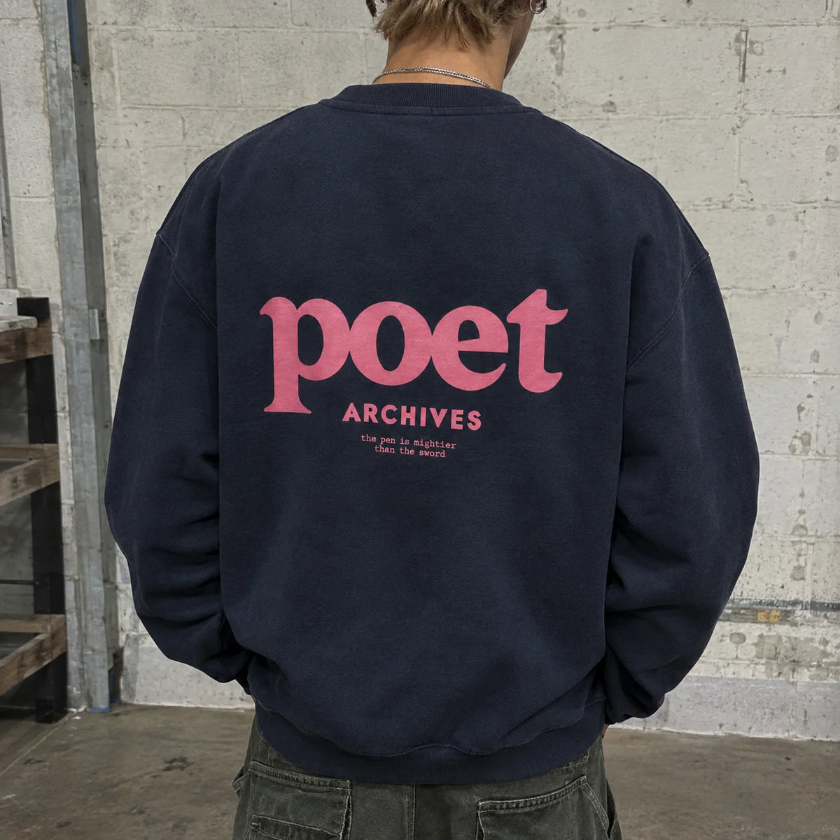 A person viewed from behind wearing a navy blue sweatshirt with a large pink graphic on the back that reads 'poet archives' and smaller text below, standing against a light concrete wall.