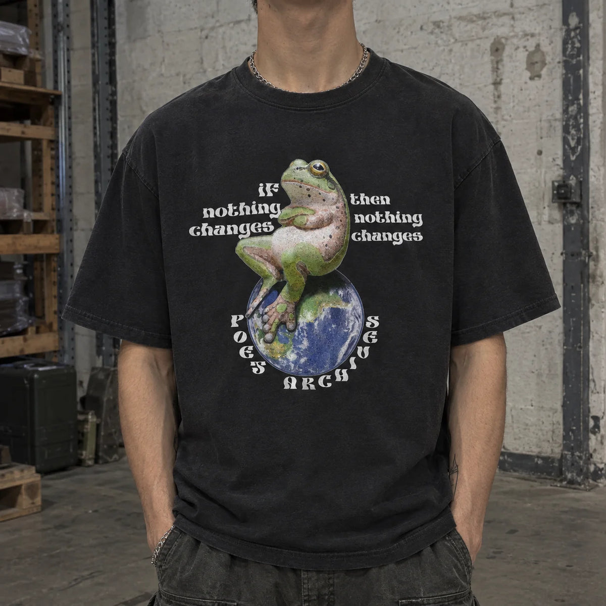 A person wearing a faded black graphic t-shirt featuring a green and pink frog sitting on a globe with the text 'IF nothing changes then nothing changes' and 'POES ARCHIVE' in a circular design. The background is a gritty, industrial warehouse setting.