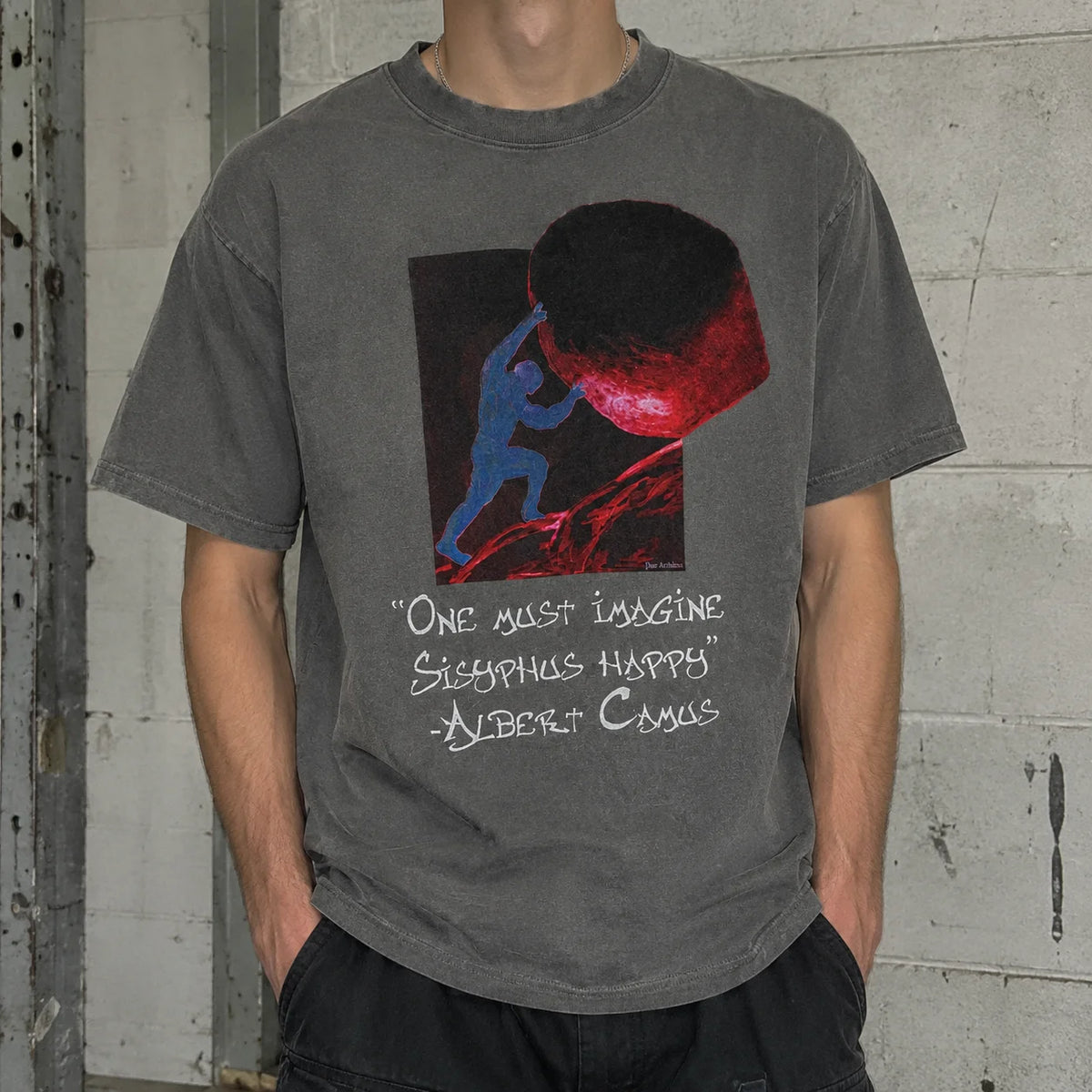 A person wearing a faded dark gray graphic t-shirt with a red and blue illustration and a quote by Albert Camus, standing against a textured gray wall.