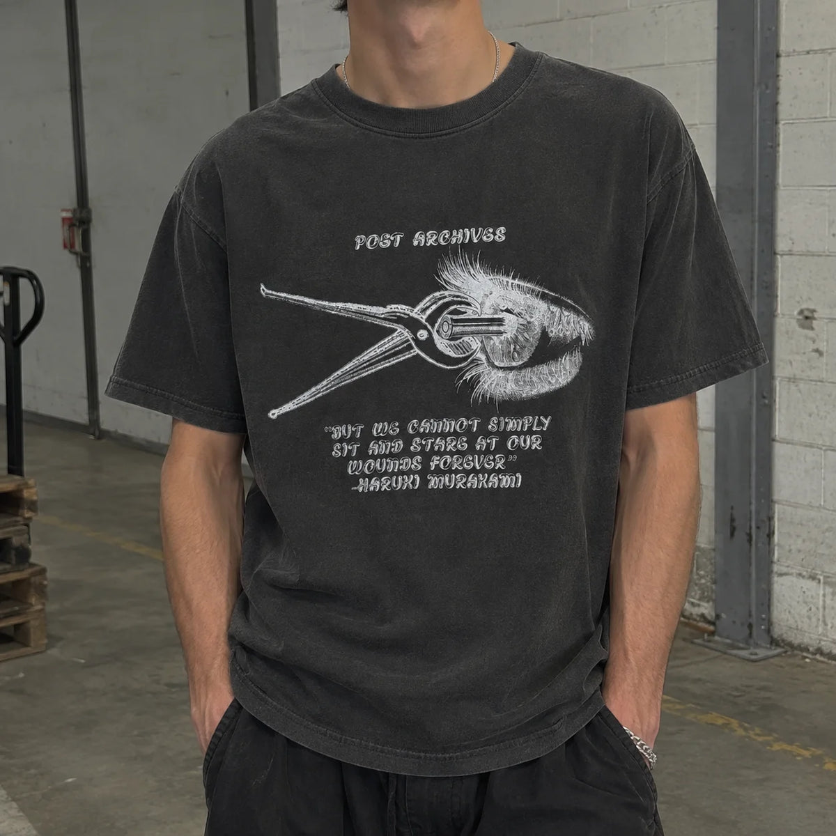 A person wearing a faded black graphic t-shirt with a white print of a hand holding a pair of shears and a quote by Haruki Murasaki. The setting is a concrete warehouse floor.